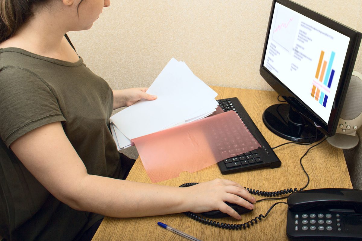 A young girl with excess weight and long hair examines documents. Work in the office. On the computer graphics monitor. Nearby phone and office supplies. Accounting, management. No dress code.