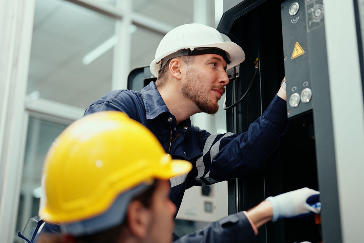 Two Caucasian production engineers in safety wear are assisting in adjusting and maintaining CNC machine in the factory. Male factory workers are examining the industrial machine to find an error.