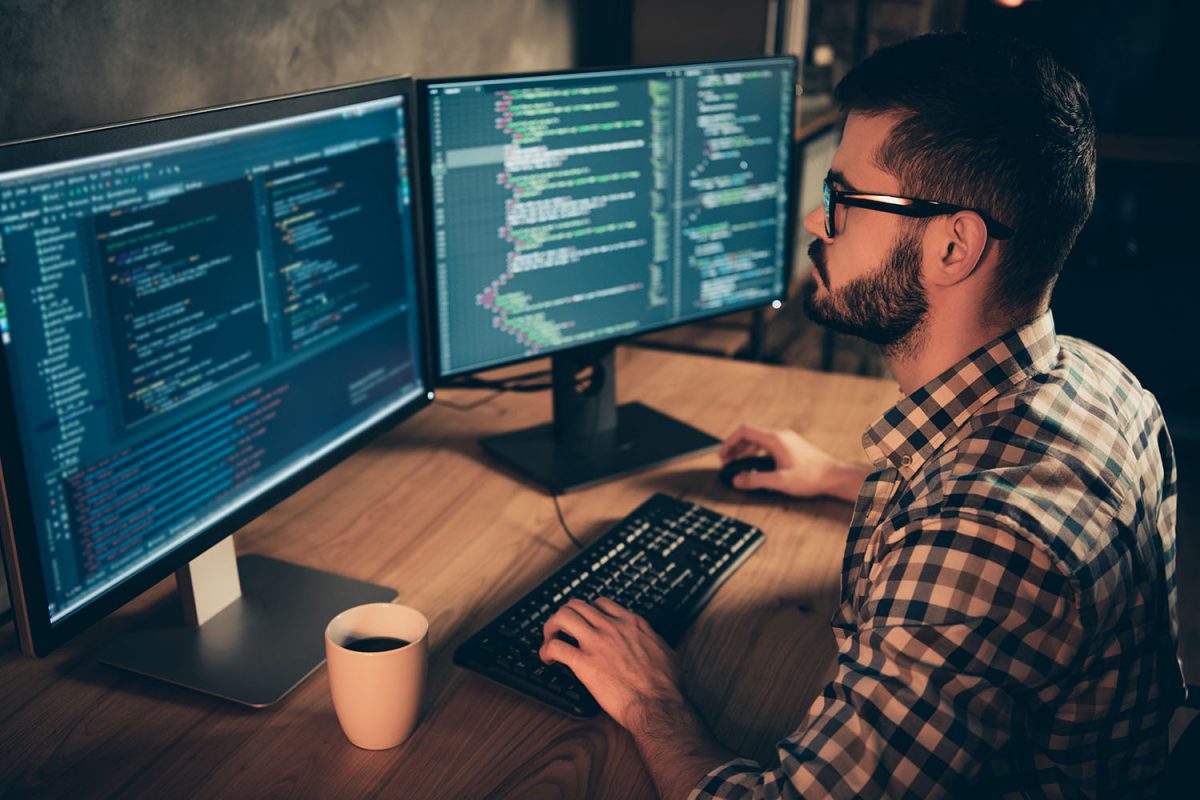 Close up side profile photo handsome he him his guy brainstorming briefing coder typing php css keyboard development outsource IT two monitors table office agency wear specs formalwear plaid shirt
